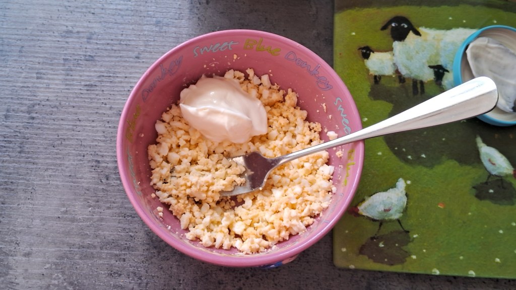 Mashed boiled egg in a bowl with a spoon of mayo, on a worktop