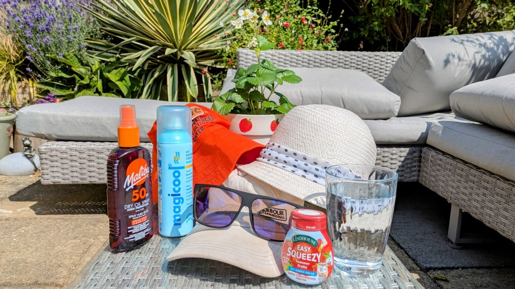 A collection of hats, a glass of water, sunscreen, cooling spray, fruit juice and sunglasses on a rattan glass table in the garden