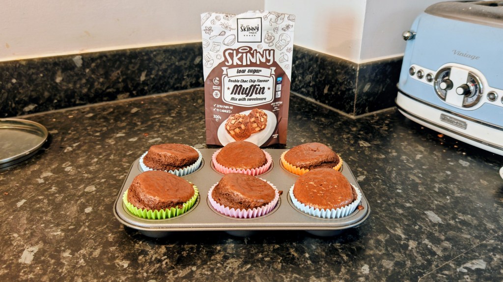 Six Skinny Food Co. Double Choc Chip Muffins in a baking tray on a worktop with the packet stood behind
