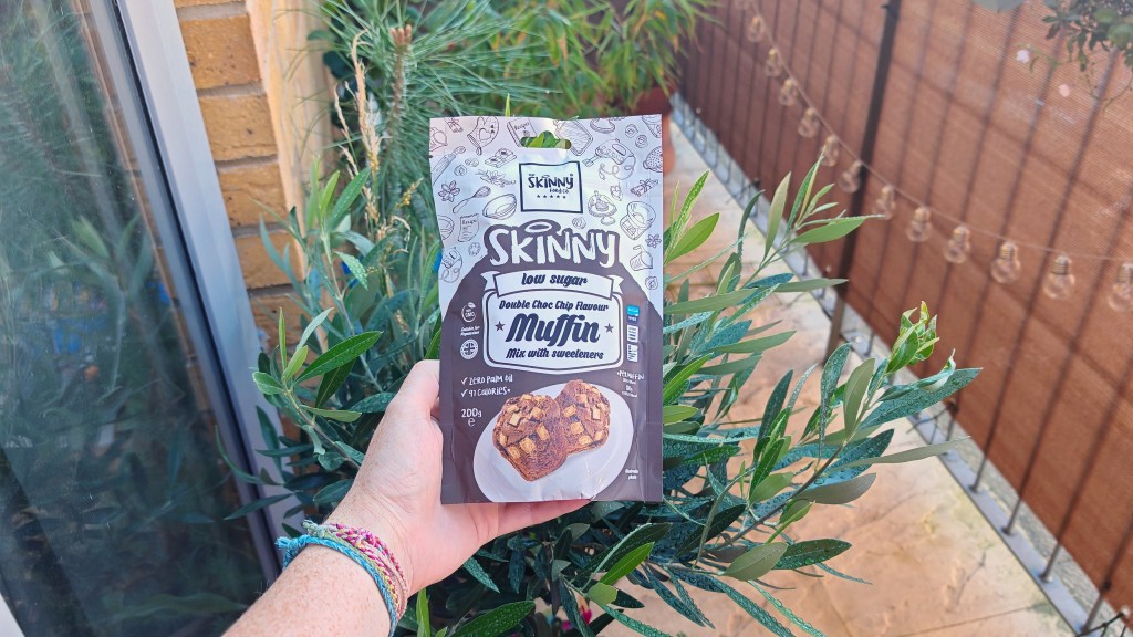 The Skinny Food Co. Double Choc Chip Muffin Mix packet being held in front of a small olive tree