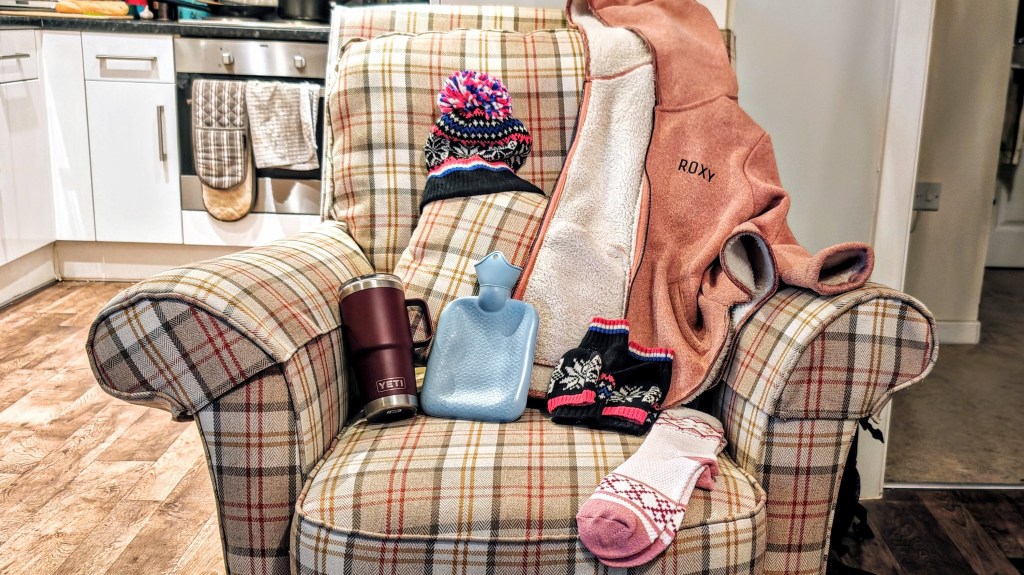 A thick fleece lined jumper, socks, hat, mittens, flask and hot water bottle on a tartan armchair
